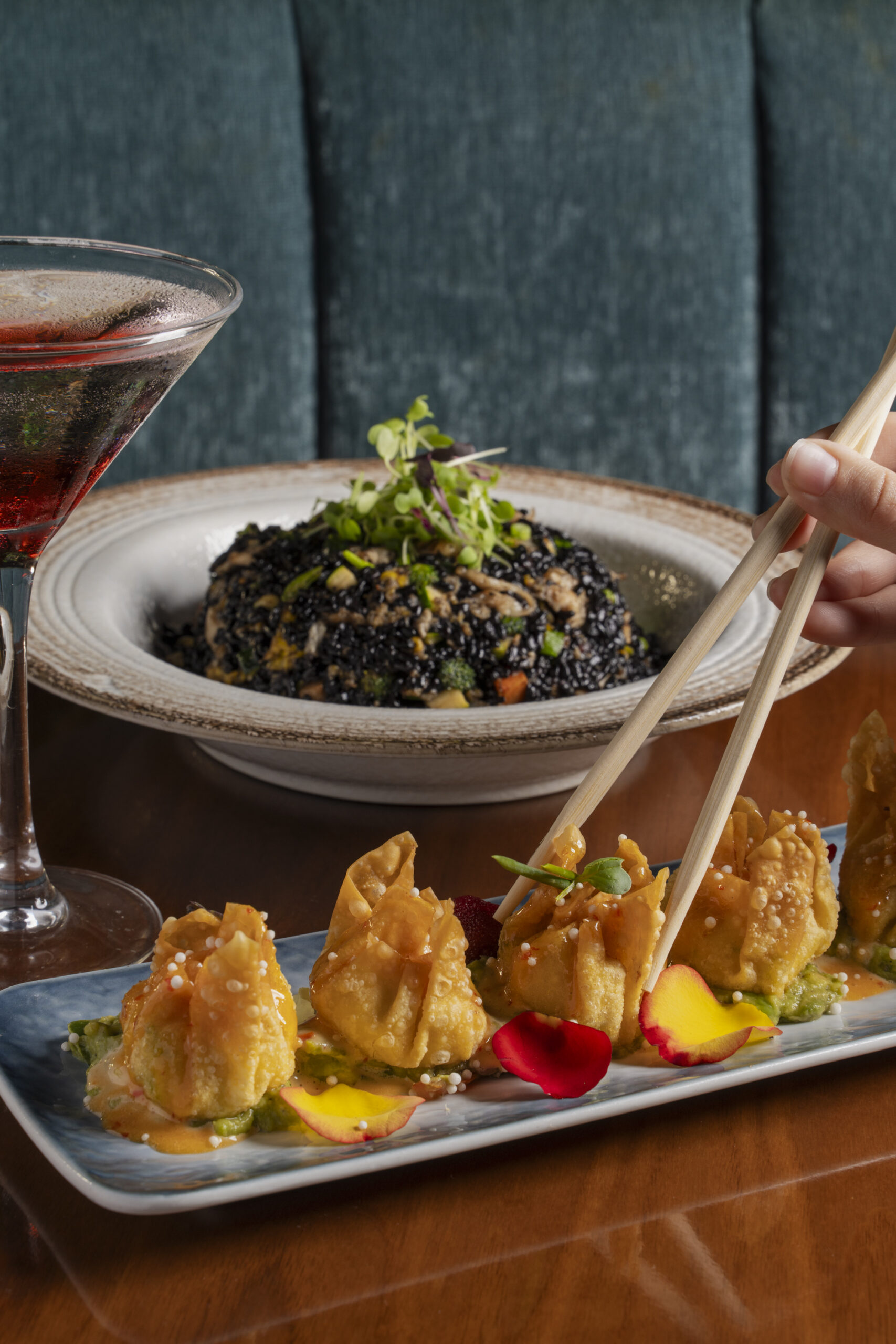 Golden-fried wontons garnished with edible flower petals on a plate, with a bowl of dark fried rice