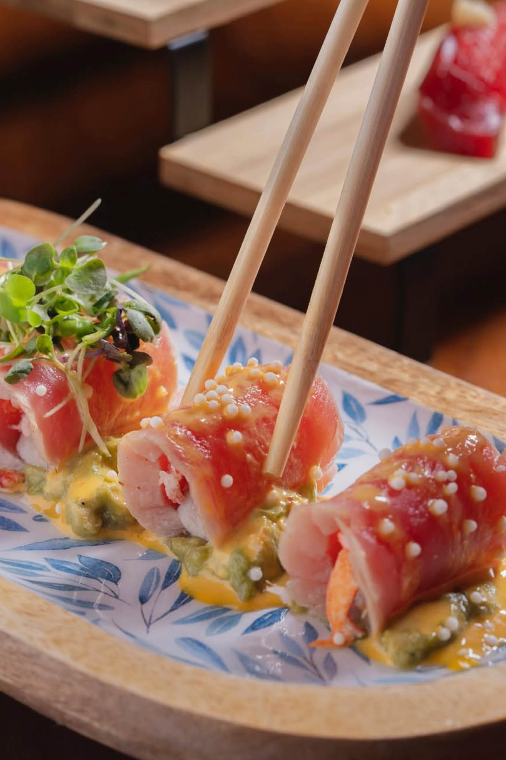 Chopsticks lifting tuna-wrapped sushi from mashed avocado and creamy sauce