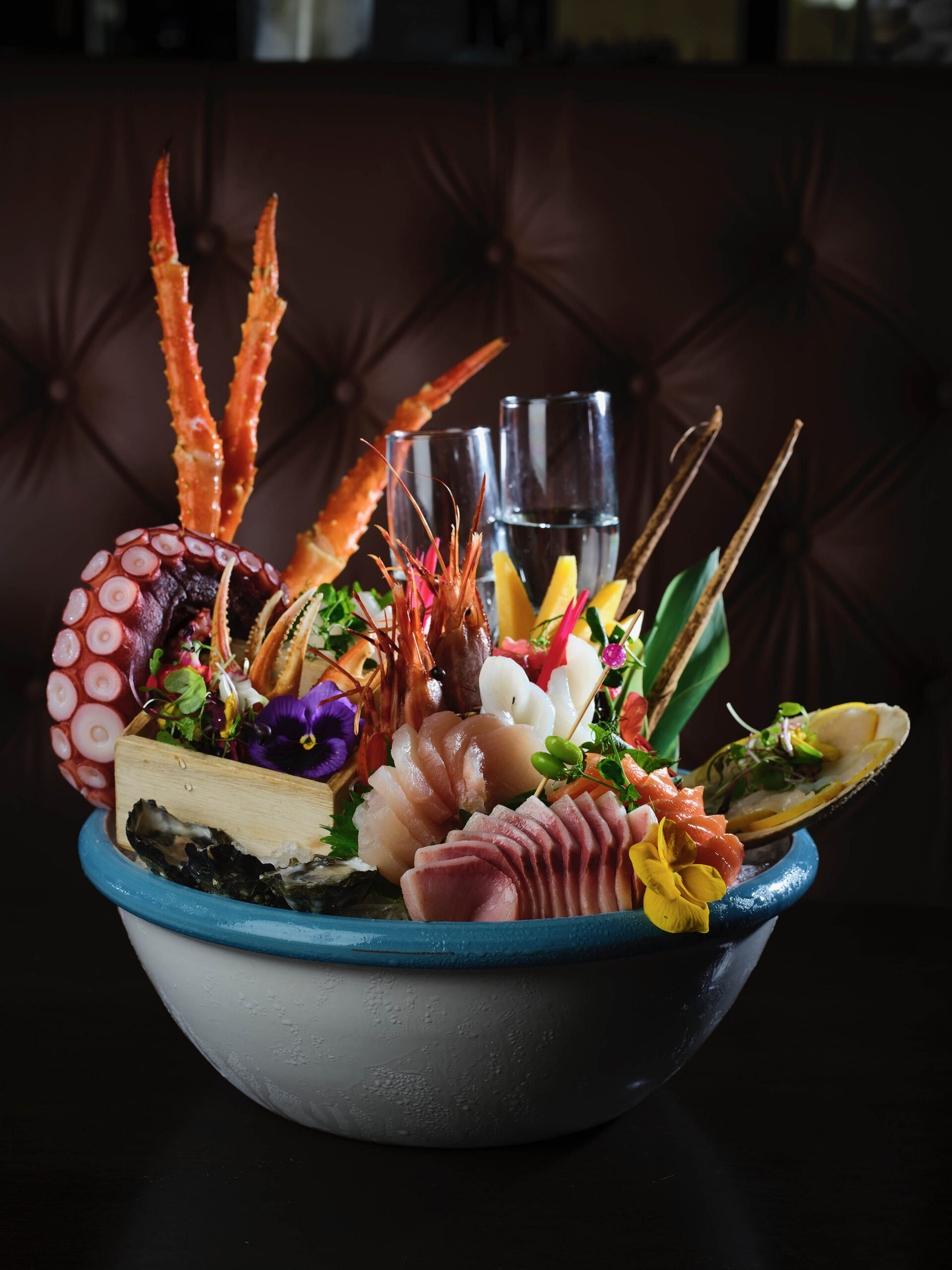 A grand seafood and sashimi platter on a table, flanked by two vibrant cocktails and three bottles of sake